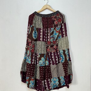Puede incluir: Falda patchwork en tonos burdeos, marrón y azul, con una variedad de estampados de cachemira y geométricos. La falda está colgada en una percha de madera contra una pared blanca. La falda tiene cintura con cordón.