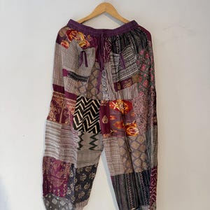 May include: Patchwork pants featuring a variety of fabric squares in shades of purple, brown, and grey. The garment has an elastic waistband with a drawstring, two front pockets with small fabric ties, and a loose, relaxed fit throughout the legs.