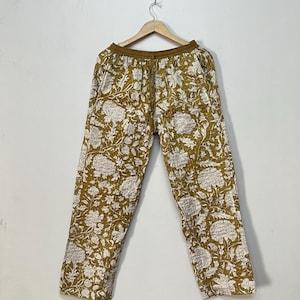 Puede incluir: Pantalones color mostaza con un estampado floral blanco, colgados de una percha de madera. Los pantalones tienen cintura con cordón y un corte relajado. El diseño floral es detallado y cubre toda la tela.
