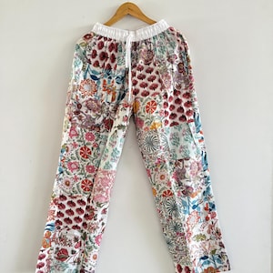 May include: Floral-print trousers in white, with a drawstring waist. The trousers have a patchwork design with various floral patterns in red, pink, orange, and blue. They are hanging on a wooden hanger against a white background.