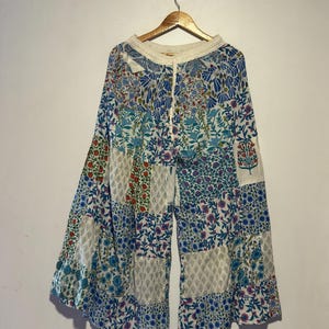Op de afbeelding: Broek met wijde pijpen en een patchwork-ontwerp. De broek heeft verschillende bloempatronen in blauw, rood en groen. De tailleband is crèmekleurig met een trekkoord. De broek hangt aan een houten hanger.