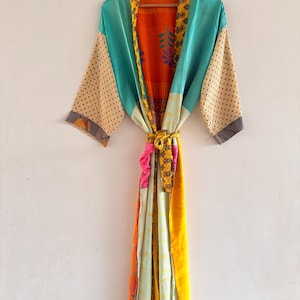 May include: A colorful, patchwork kimono robe with a tie belt, hanging on a wooden hanger. The robe features panels of teal, orange, yellow, and beige with polka dots and floral patterns. The robe is made of a lightweight fabric.