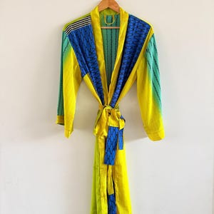 Puede incluir: Una bata de manga larga con un diseño de patchwork vibrante en amarillo brillante, azul verdoso y azul. La prenda incluye un cinturón a juego en la cintura, un cuello tipo chal y paneles en contraste con patrones geométricos y rayas verticales.