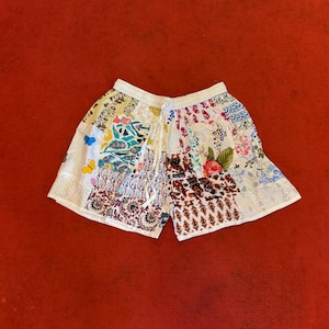 Può includere: Pantaloncini bianchi patchwork con vita con coulisse. I pantaloncini presentano varie toppe di tessuto a motivi floreali nei toni del blu, rosa, verde e viola. Una farfalla gialla è visibile su una delle toppe. I pantaloncini sono appoggiati su una superficie rossa.