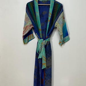 May include: A long-sleeved kimono robe featuring a patchwork design of blue, teal, and green fabric panels. The garment includes a matching tie belt at the waist and a wide collar, showcasing various geometric and floral patterns throughout the fabric.