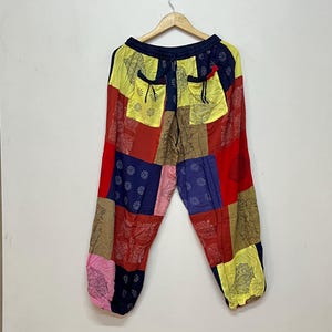 Op de afbeelding: Patchwork broek met een levendig kleurenpalet van rood, geel, blauw, roze en beige. De broek heeft een elastische tailleband met een trekkoord, twee voorzakken en is versierd met diverse zwarte bloem- en zonachtige patronen.