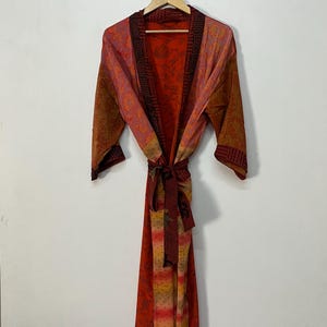Könnte beinhalten: Ein langer, fließender Bademantel mit einem lebendigen, gemusterten Design. Der Bademantel weist eine Mischung aus warmen Farben auf, darunter Rot, Orange und Braun, mit komplizierten Details. Er hängt an einem Holzbügel und zeigt seine volle Länge und sein Design.