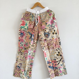 May include: Patchwork pants with a floral design in shades of pink, beige, and blue. The pants have a white drawstring waistband and are hanging on a wooden hanger against a white background. The fabric appears to be lightweight.