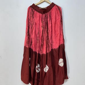 Puede incluir: Falda larga y fluida en tonos rojos y burdeos, colgada de una percha de madera. La falda presenta un estampado tie-dye con detalles blancos cerca del dobladillo. La tela parece ligera y adecuada para el clima cálido.