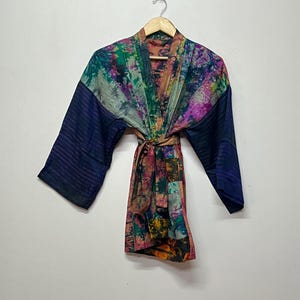 Könnte beinhalten: Ein farbenfroher Kimono-Bademantel mit Muster und langen Ärmeln. Der Bademantel weist eine Mischung aus leuchtenden Farben auf, darunter Blau, Grün, Rosa und Lila. Die Ärmel sind einfarbig Marineblau mit einem Streifenmuster. Der Bademantel wird mit einem passenden Gürtel in der Taille gebunden.