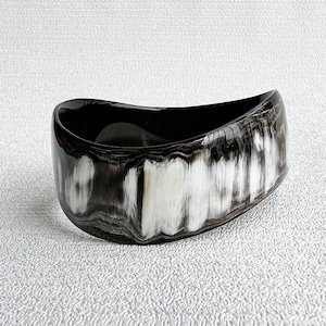 Pu&ograve; includere: Un bracciale rigido largo e curvo con finitura lucida e un motivo marmorizzato di venature nero profondo, grigio antracite e bianco crema. Il bracciale ha una forma organica asimmetrica con una superficie liscia e brillante.