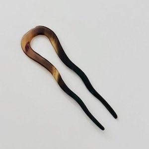 Può includere: Una forcina per capelli con una parte superiore curva a forma di U e due lunghe e sottili punte. La parte superiore è marrone marmorizzato, che sfuma in nero alle punte. L'accessorio per capelli è posto su uno sfondo bianco.