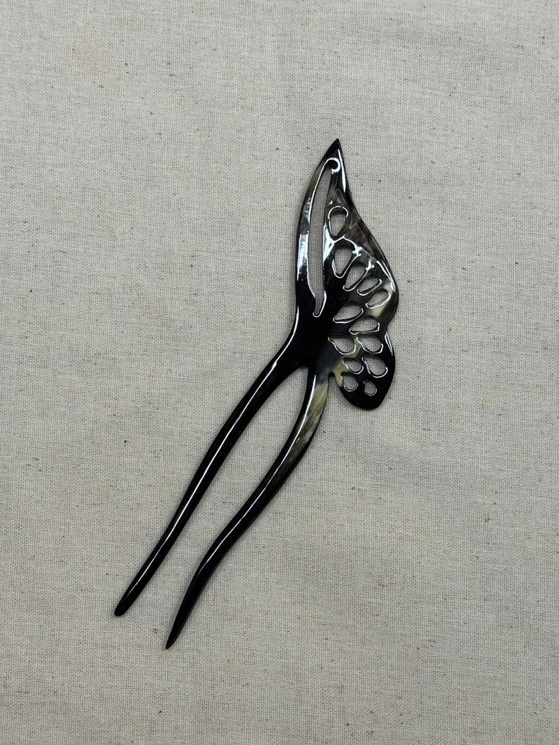 May include: A black butterfly-shaped hair fork with a glossy finish. The butterfly wing design features cut-out details. The hair accessory has two long, slender prongs extending from the wing.