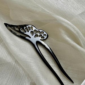 May include: A black and white hair fork with a decorative openwork design. The hair accessory has two long, pointed prongs and a curved top with a series of circular cutouts. The hair fork is set against a white, textured fabric.