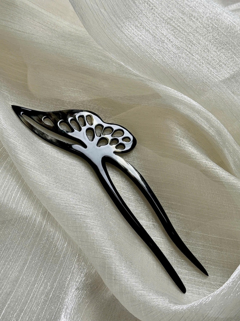 May include: A black and white hair fork with a decorative openwork design. The hair accessory has two long, pointed prongs and a curved top with a series of circular cutouts. The hair fork is set against a white, textured fabric.