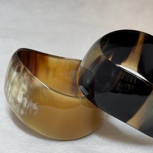 Pu&ograve; includere: Due bracciali rigidi larghi con una finitura liscia e lucida. Uno presenta un motivo marmorizzato traslucido color crema e marrone chiaro, mentre l'altro mostra una sfumatura nero profondo e marrone scuro con una superficie lucida e riflettente.