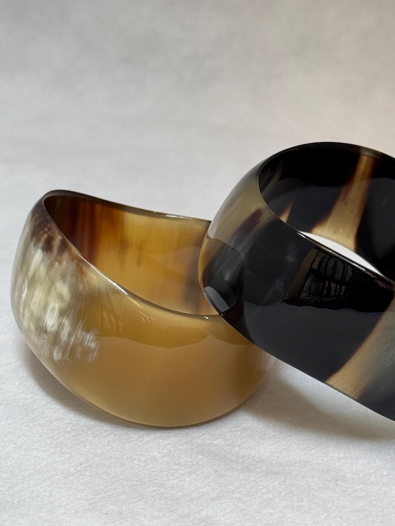Pu&ograve; includere: Due bracciali rigidi larghi con una finitura liscia e lucida. Uno presenta un motivo marmorizzato traslucido color crema e marrone chiaro, mentre l'altro mostra una sfumatura nero profondo e marrone scuro con una superficie lucida e riflettente.