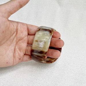 Pu&ograve; includere: Un bracciale rigido largo realizzato in corno lucidato, caratterizzato da una superficie liscia e arrotondata con un motivo marmorizzato naturale di striature traslucide color crema, beige e marrone scuro che percorrono tutto il materiale.