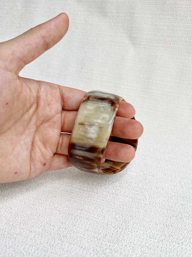 Pu&ograve; includere: Un bracciale rigido largo realizzato in corno lucidato, caratterizzato da una superficie liscia e arrotondata con un motivo marmorizzato naturale di striature traslucide color crema, beige e marrone scuro che percorrono tutto il materiale.