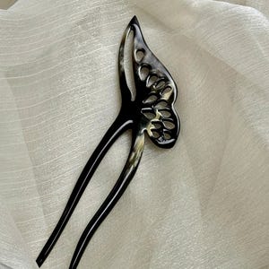 May include: A black and cream-colored butterfly hair stick. The hair accessory features a butterfly wing design with cut-out details and two long, slender prongs. The hair stick is set against a white, textured background.