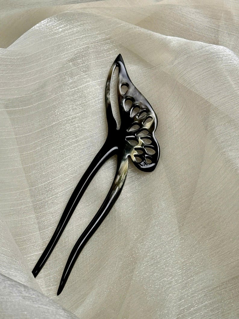May include: A black and cream-colored butterfly hair stick. The hair accessory features a butterfly wing design with cut-out details and two long, slender prongs. The hair stick is set against a white, textured background.