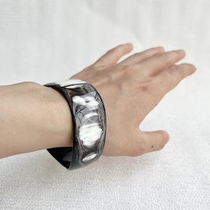 Pu&ograve; includere: Un bracciale rigido largo e lucido caratterizzato da un motivo marmorizzato con venature nero profondo e bianco traslucido. La superficie liscia e curva del bracciale presenta una finitura lucida con variazioni di colore organiche e vorticose.