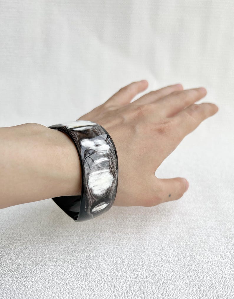 Pu&ograve; includere: Un bracciale rigido largo e lucido caratterizzato da un motivo marmorizzato con venature nero profondo e bianco traslucido. La superficie liscia e curva del bracciale presenta una finitura lucida con variazioni di colore organiche e vorticose.