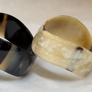 Pu&ograve; includere: Due bracciali rigidi larghi con una finitura liscia e lucida. Un bracciale &egrave; principalmente grigio antracite e nero con sottili venature verticali, mentre l'altro &egrave; color crema e beige traslucido con delicate inclusioni marmorizzate marroni.