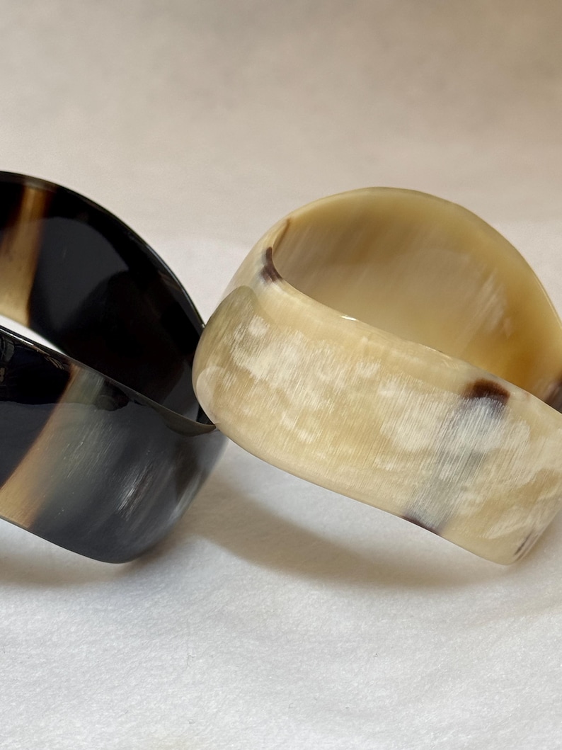 Pu&ograve; includere: Due bracciali rigidi larghi con una finitura liscia e lucida. Un bracciale &egrave; principalmente grigio antracite e nero con sottili venature verticali, mentre l'altro &egrave; color crema e beige traslucido con delicate inclusioni marmorizzate marroni.
