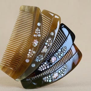May include: A set of four hair combs in varying shades of brown and black, each featuring an inlaid mother-of-pearl floral and butterfly design. The combs have smooth, curved edges and fine teeth, showcasing natural horn-like patterns and textures.