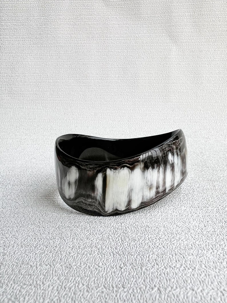 Pu&ograve; includere: Un bracciale rigido largo e curvo con finitura lucida e un motivo marmorizzato di venature nero profondo, grigio antracite e bianco crema. Il bracciale ha una forma organica asimmetrica con una superficie liscia e brillante.