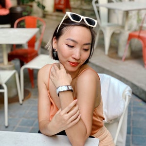 Pu&ograve; includere: Un ampio bracciale a fascia metallico con una superficie iridescente e riflettente indossato al polso. Il bracciale presenta un design liscio e curvo che cattura la luce, abbinato a piccoli orecchini pendenti triangolari scuri e occhiali da sole con montatura bianca.