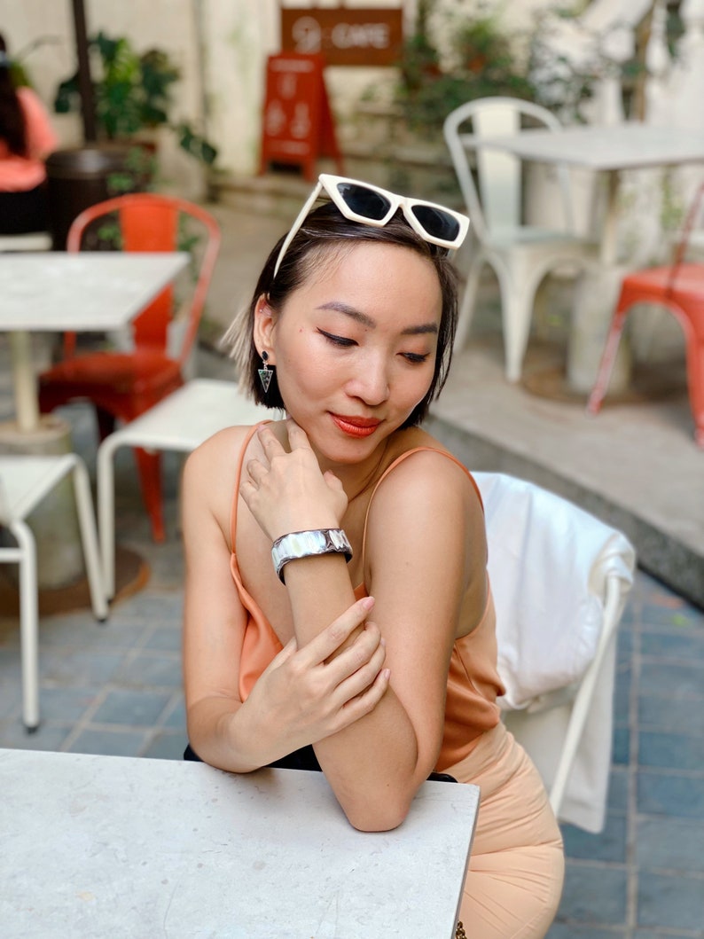 Pu&ograve; includere: Un ampio bracciale a fascia metallico con una superficie iridescente e riflettente indossato al polso. Il bracciale presenta un design liscio e curvo che cattura la luce, abbinato a piccoli orecchini pendenti triangolari scuri e occhiali da sole con montatura bianca.