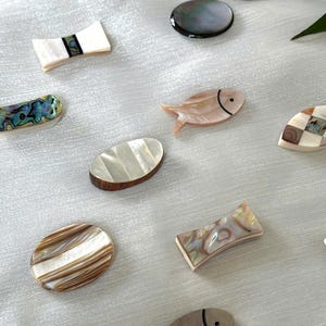 Puede incluir: Una colección de objetos decorativos, incluyendo formas ovaladas y rectangulares, y piezas con forma de pez. Los artículos presentan patrones iridiscentes y naturales de concha en tonos blancos, marrones y rosas. El logotipo de Emoi Hanoi es visible.