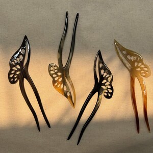 May include: Four hair forks with butterfly wing designs. Two are black, one is a light brown, and one is a mix of light and dark brown. The butterfly wings have cut-out patterns. The forks are on a neutral-colored background.