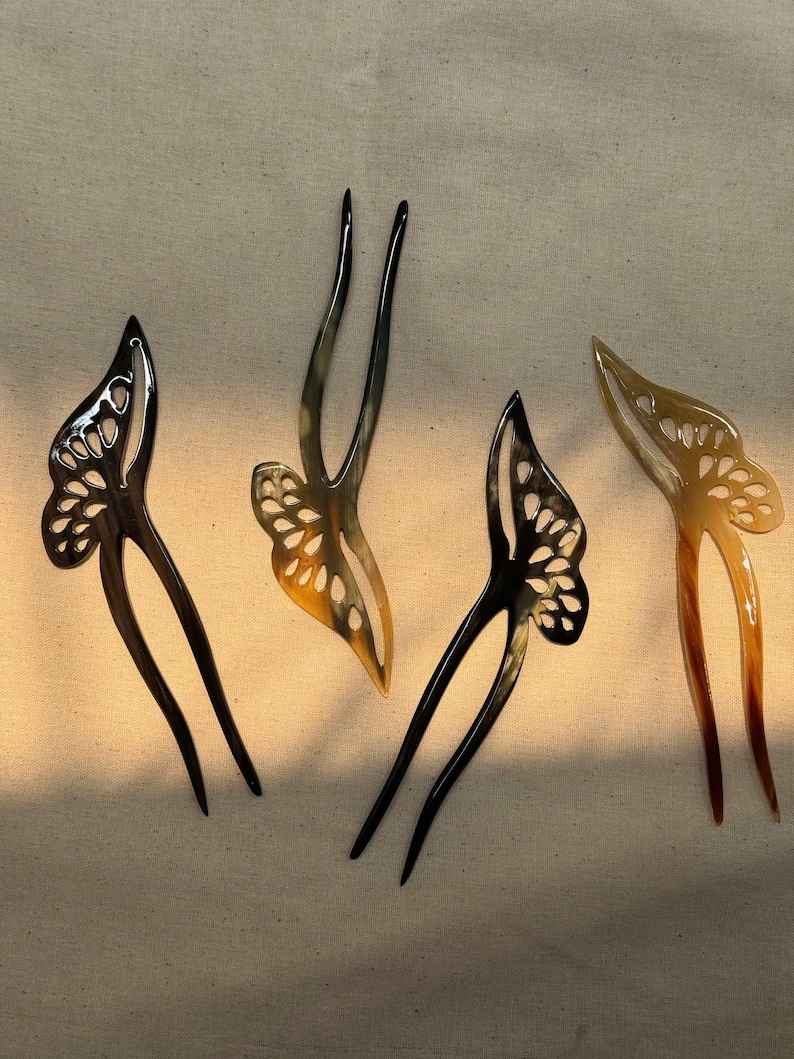 May include: Four hair forks with butterfly wing designs. Two are black, one is a light brown, and one is a mix of light and dark brown. The butterfly wings have cut-out patterns. The forks are on a neutral-colored background.