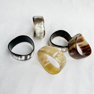 Pu&ograve; includere: Una collezione di cinque anelli lisci e levigati dalle forme organiche irregolari. Gli anelli presentano vari motivi naturali nelle tonalit&agrave; crema, beige, marrone scuro e nero, mostrando un aspetto traslucido simile al corno con sfumature di colore uniche.
