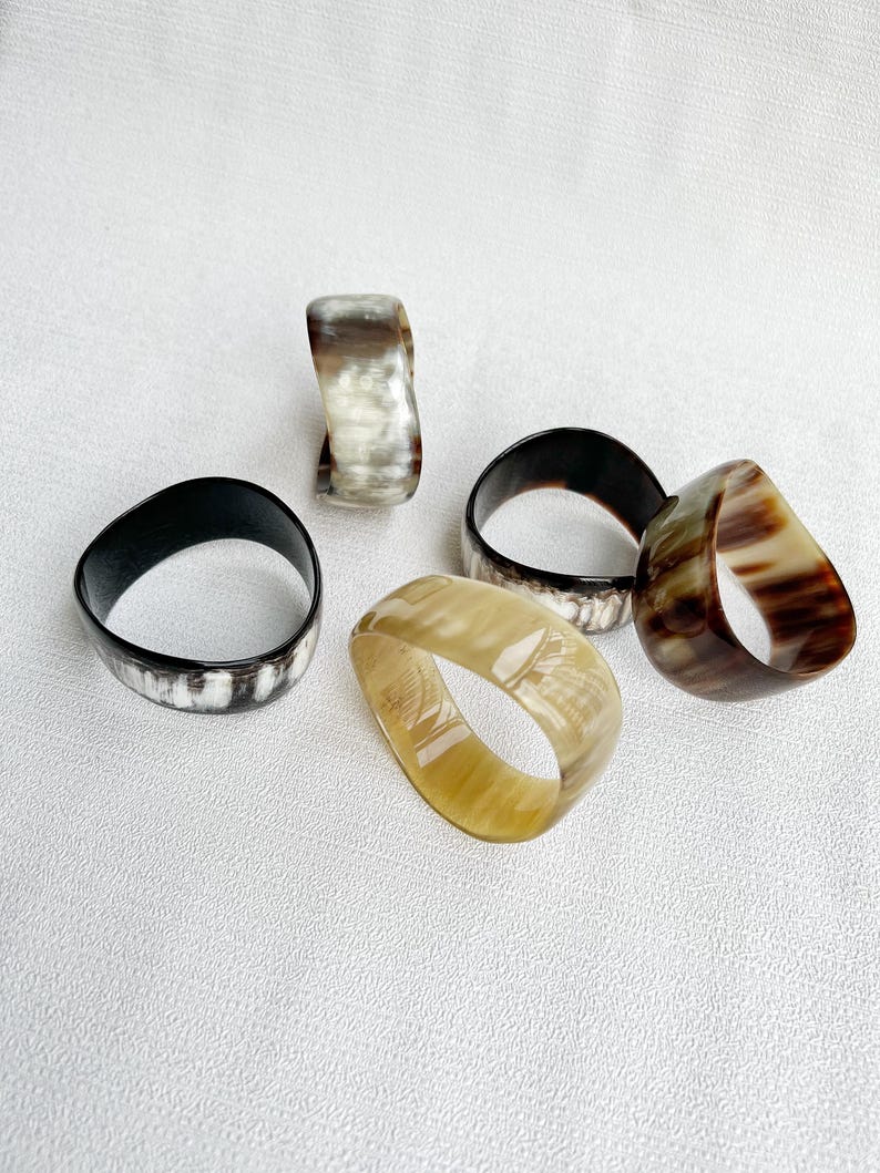 Pu&ograve; includere: Una collezione di cinque anelli lisci e levigati dalle forme organiche irregolari. Gli anelli presentano vari motivi naturali nelle tonalit&agrave; crema, beige, marrone scuro e nero, mostrando un aspetto traslucido simile al corno con sfumature di colore uniche.