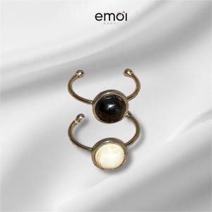 Puede incluir: Dos anillos plateados con piedras circulares negras y blancas. Cada anillo presenta una banda curva con una pequeña bola en cada extremo y una piedra central. Los anillos se muestran sobre una superficie blanca texturizada. La marca "emoi hanoi" está en la esquina superior izquierda.