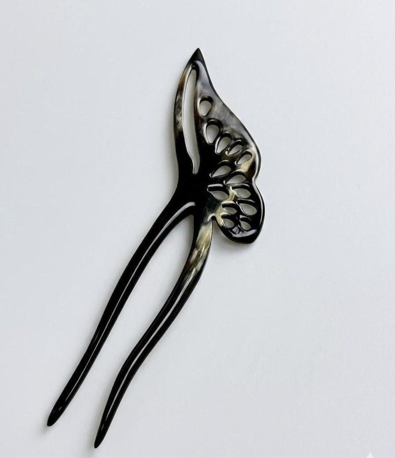 May include: A black and cream-colored hair fork with a butterfly wing design. The hair accessory has a glossy finish and a forked base. The butterfly wing features cut-out details.