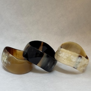 Pu&ograve; includere: Tre bracciali rigidi larghi realizzati in corno lucidato. Il bracciale a sinistra &egrave; di un colore beige traslucido, quello centrale &egrave; nero intenso con sottili venature grigie e quello a destra &egrave; bianco crema con motivi maculati marrone scuro.