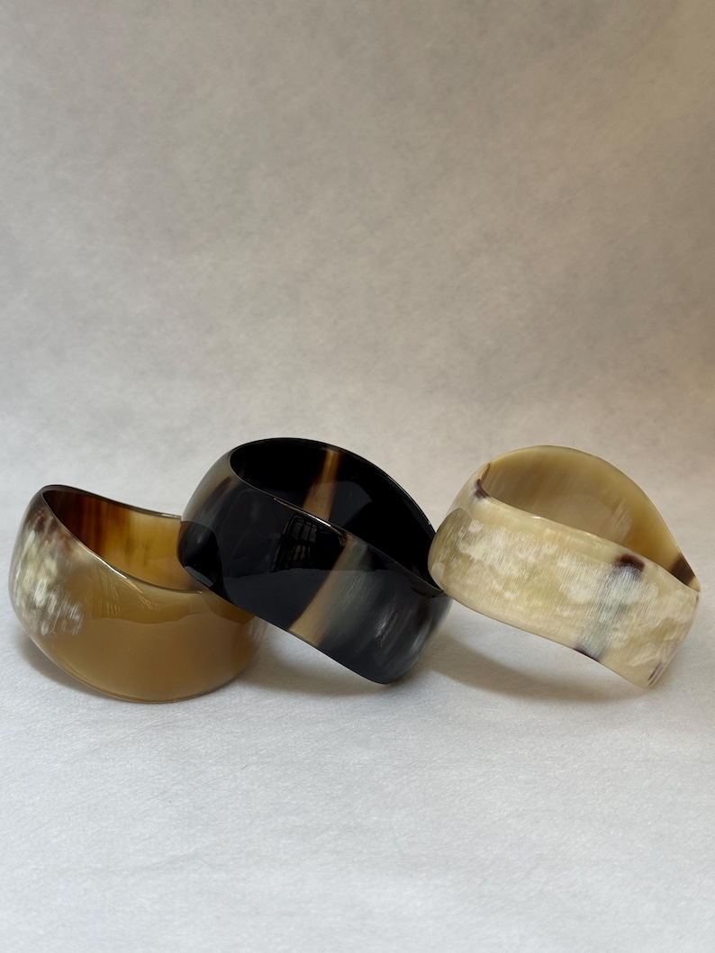 Pu&ograve; includere: Tre bracciali rigidi larghi realizzati in corno lucidato. Il bracciale a sinistra &egrave; di un colore beige traslucido, quello centrale &egrave; nero intenso con sottili venature grigie e quello a destra &egrave; bianco crema con motivi maculati marrone scuro.