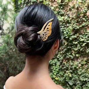 May include: A decorative butterfly hair clip in shades of cream and gold is placed in dark hair styled in a bun. The clip is made of a light-colored material and has intricate cut-out details. The background features green foliage.