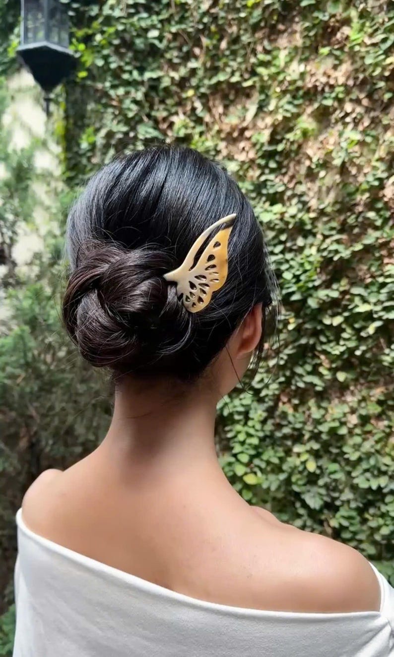 May include: A decorative butterfly hair clip in shades of cream and gold is placed in dark hair styled in a bun. The clip is made of a light-colored material and has intricate cut-out details. The background features green foliage.