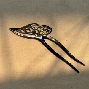 May include: A dark, glossy hair pin with a decorative, openwork design. The pin features a leaf-like shape with a series of cut-out circles at the top and two long, slender prongs. The hair accessory is made of a dark material with a subtle, natural pattern.
