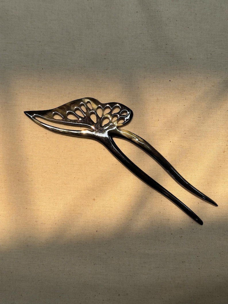 May include: A dark, glossy hair pin with a decorative, openwork design. The pin features a leaf-like shape with a series of cut-out circles at the top and two long, slender prongs. The hair accessory is made of a dark material with a subtle, natural pattern.