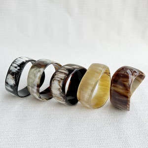 Pu&ograve; includere: Cinque bracciali rigidi lisci e lucidi disposti in fila. Ogni bracciale presenta un motivo naturale unico con varie tonalit&agrave; di nero, bianco, grigio, marrone chiaro e marrone scuro, con una finitura lucida e semitrasparente.
