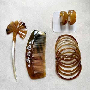 May include: A collection of hair accessories and jewelry in shades of brown. The items include a hair comb, a hair stick, hoop earrings, and several bangles. The comb is decorated with silver-colored floral accents. The earrings are on a white card.