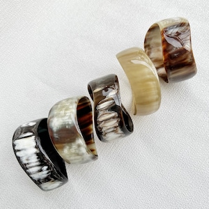 Pu&ograve; includere: Cinque bracciali a fascia larghi e lucidi disposti in linea diagonale. Ogni bracciale presenta un motivo organico unico con varie tonalit&agrave; di marrone, crema, nero e beige traslucido, con una superficie liscia, lucida e curva.