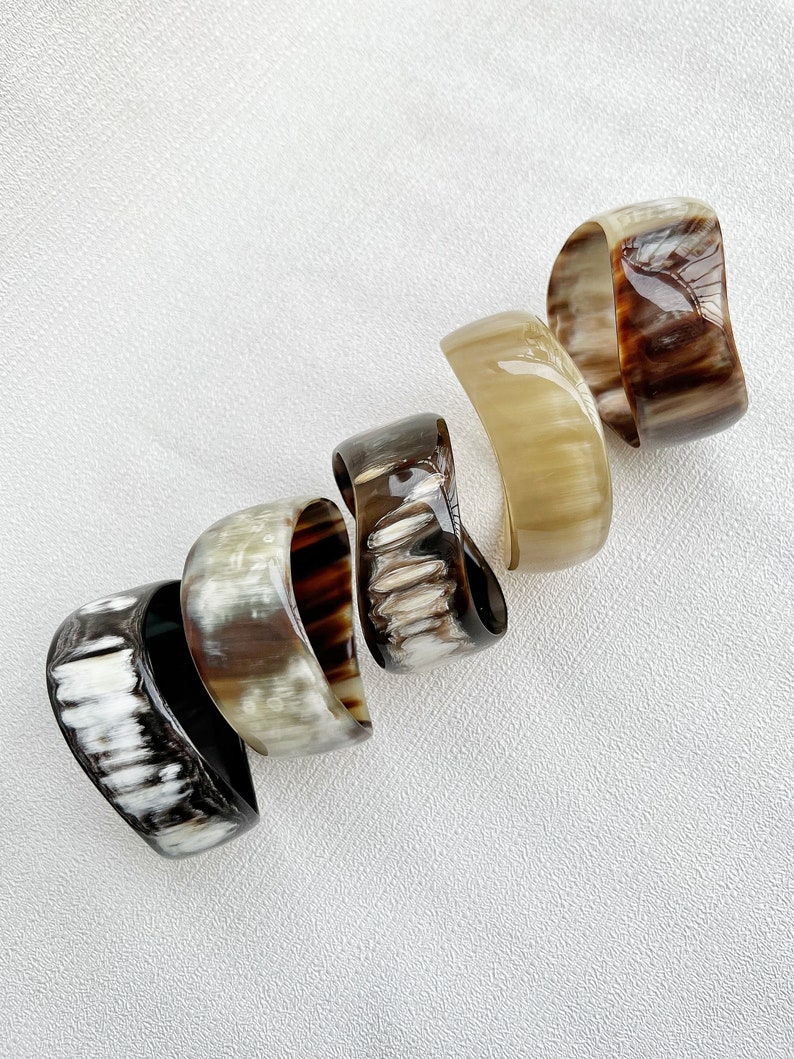 Pu&ograve; includere: Cinque bracciali a fascia larghi e lucidi disposti in linea diagonale. Ogni bracciale presenta un motivo organico unico con varie tonalit&agrave; di marrone, crema, nero e beige traslucido, con una superficie liscia, lucida e curva.